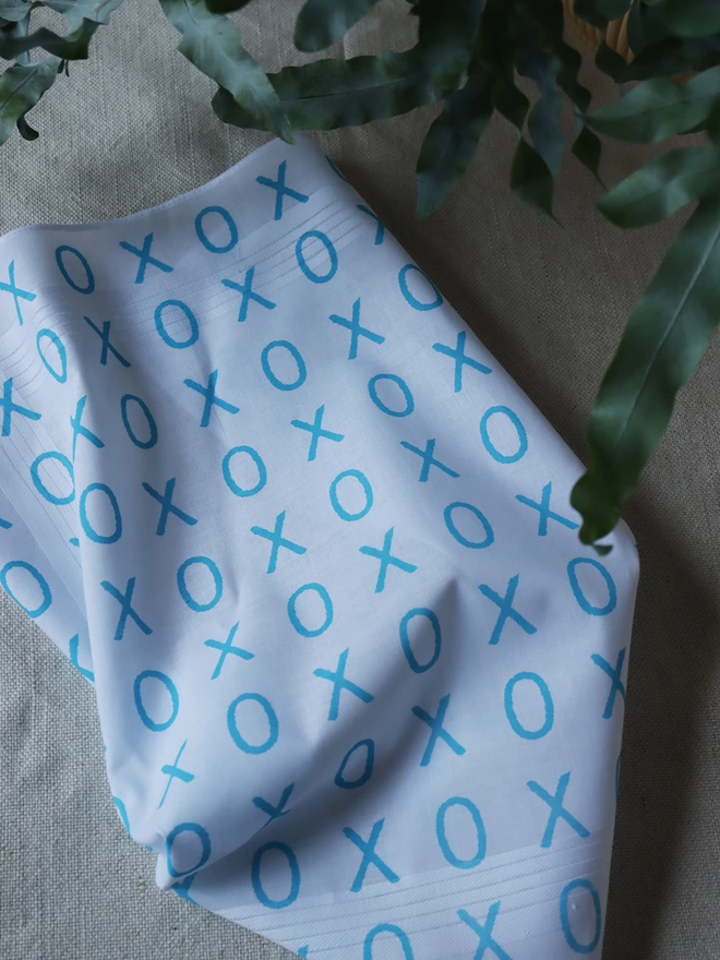 A Mr.PS Hugs and Kisses handkerchief printed in aqua on a linen tablecloth with leaves of a houseplant