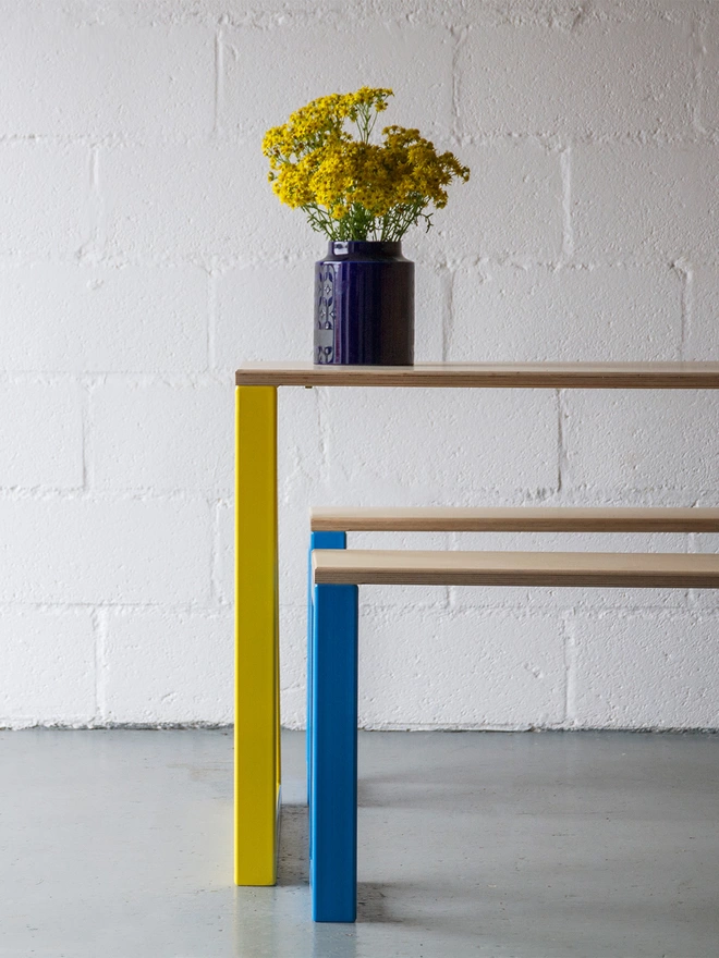 end view of chunky blue bench frame and yellow table frame of a contemporary nola dining set with a blue vase of yellow flowers on top