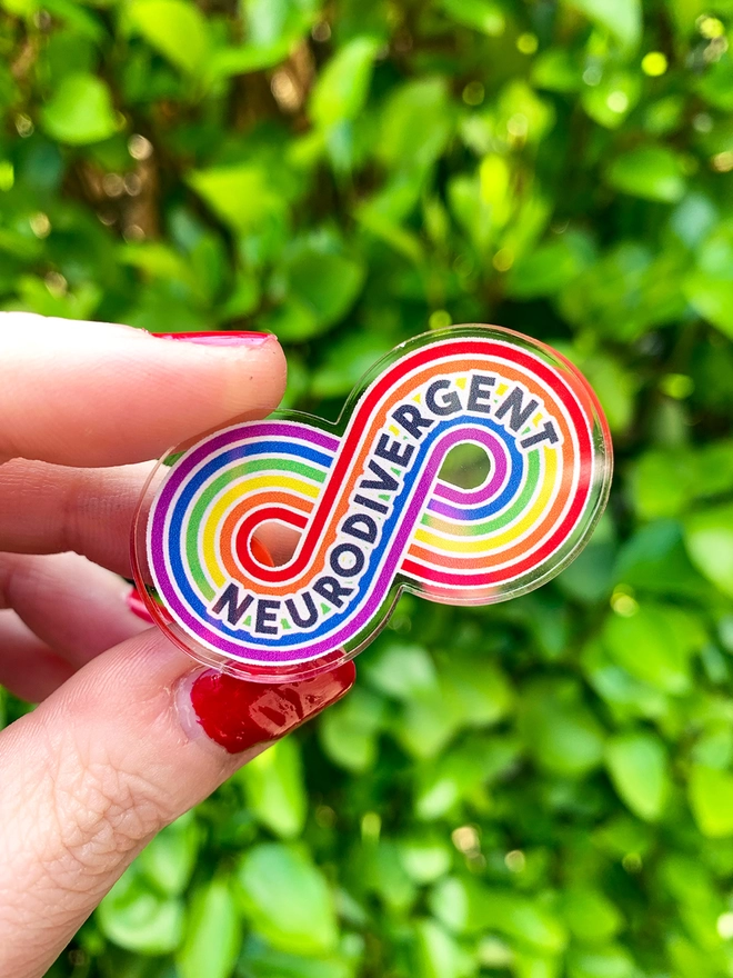 image shows a hand holding an acrylic badge in the shape of a rainbow stripe infinity loop. printed across the loop is the word 'Neurodivergent'. 