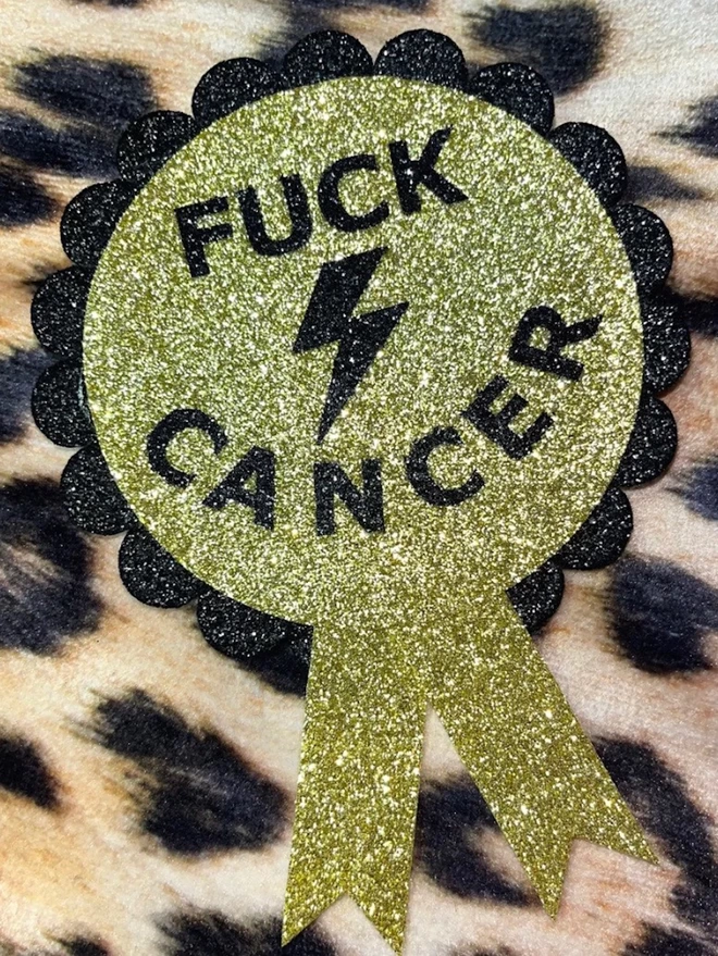  A rosette with a gold disc in the centre featuring the words ‘FUCK CANCER’ and a lightening bolt in black. The scalloped edging is black. The tails are gold.
