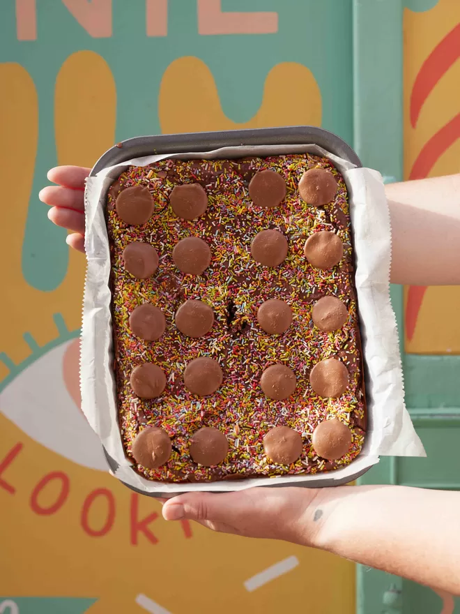 Freshly baked Funfetti brownies with sprinkles and milk chocolate buttons being held by two hands in a baking tray against a colourful background