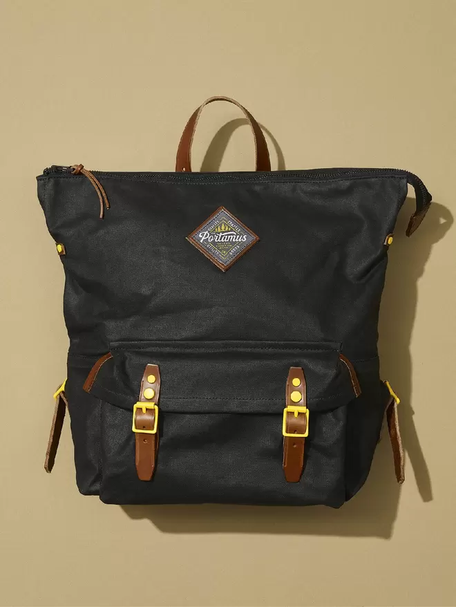 Black zip top backpack with brown leather trim and yellow hardware on taupe background.
