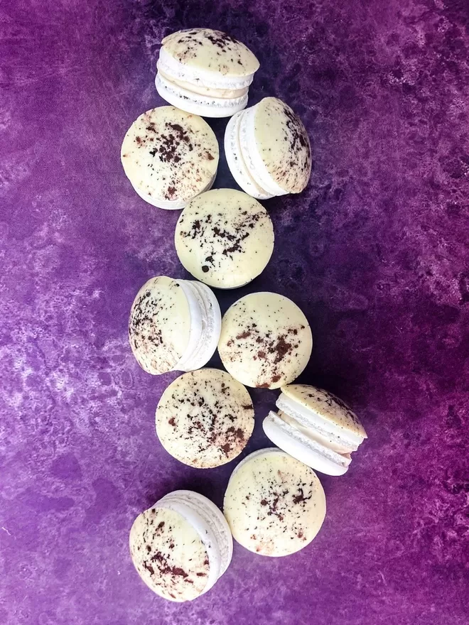 several white macarons with a white chocolate disc and coco powder dusted on top, piled on a purple background