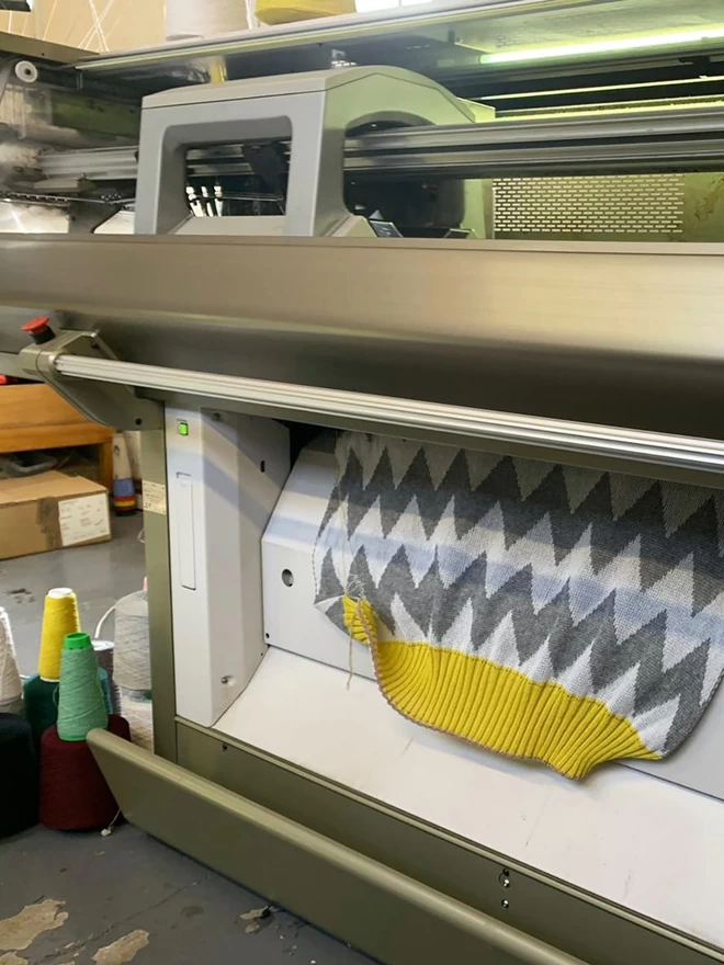 A chevron blanket being knitted on an industrial Shima Seiki knitting machine. The grey and white zigzag fabric with a mustard yellow trim can be seen coming out of the machine and several colourful cones of yarn are situated on the floor.
