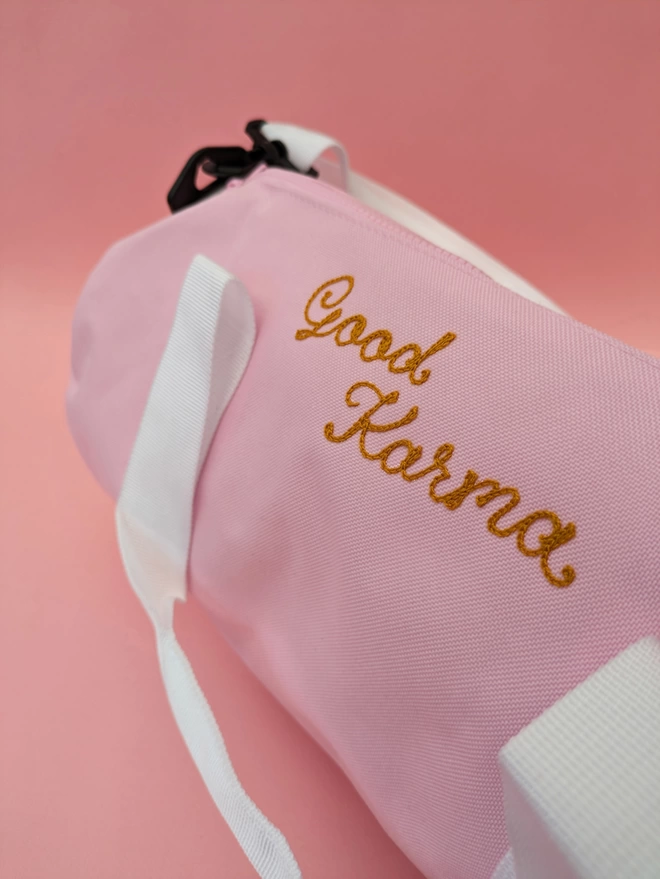 Pink Duffel Bag close up detail with white straps and handles, personalised with gold embroidery reading 'Good Karma' on a pink background