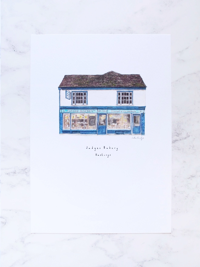 Watercolour illustration of Judges Bakery a beautiful blue shopfront with windows full of bakery goods sits below a white first floor and dark brown / grey roof.  The illustration is a small watercolour painting in the centre of the white page, the photo background is a pale white marble.