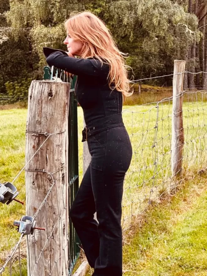 Patti Jumpsuit In Denim Black model standing leaning against a fence