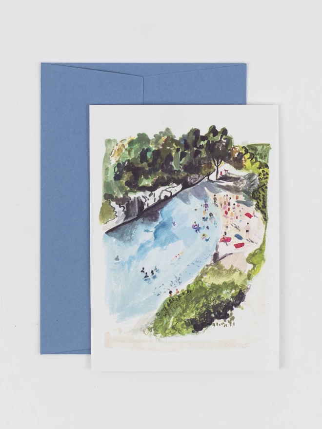 A greetings card featuring an observational drawing of a rocky beach in France. The small cove is full of people sunbathing and surrounded by trees. There are a few people in the water swimming. With a corresponding pale blue envelope.