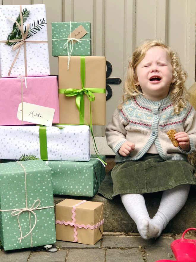 A stack of gifts wrapped in green and white paper with a tiny tree design are piled beside a toddler.