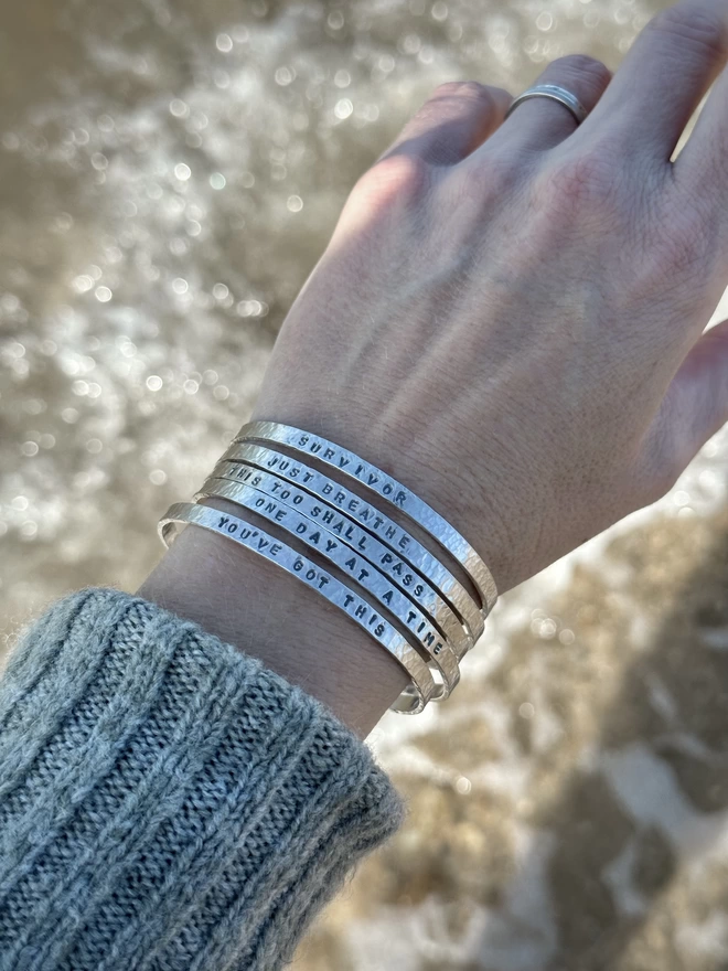 The left wrist of a woman in a knitted green jumper, wearing a stack of five sterling silver cuff bangles  with stamped text, above the surf of an ocean shoreline.