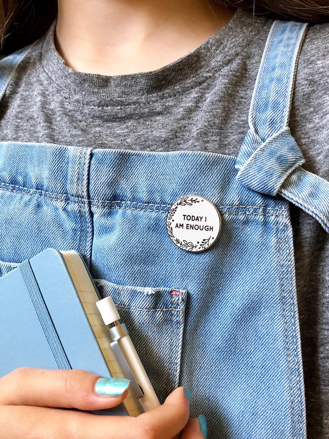 A white enamel pin is pinned to someone's blue dungarees. The pin is round with a botanical design and the words "Today I Am Enough".
