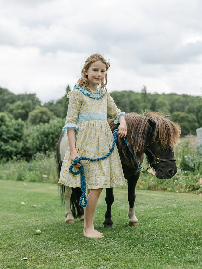 A girl stands with a miniature pony. She is wearing a yellow floral dress with a blue frill collar and sleeves and a blue sash