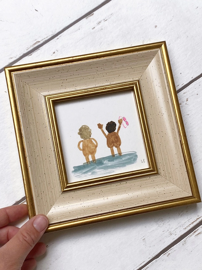 hand holding framed picture of two naked people cheerfully looking out to sea with pink goggles in cream and gold frame