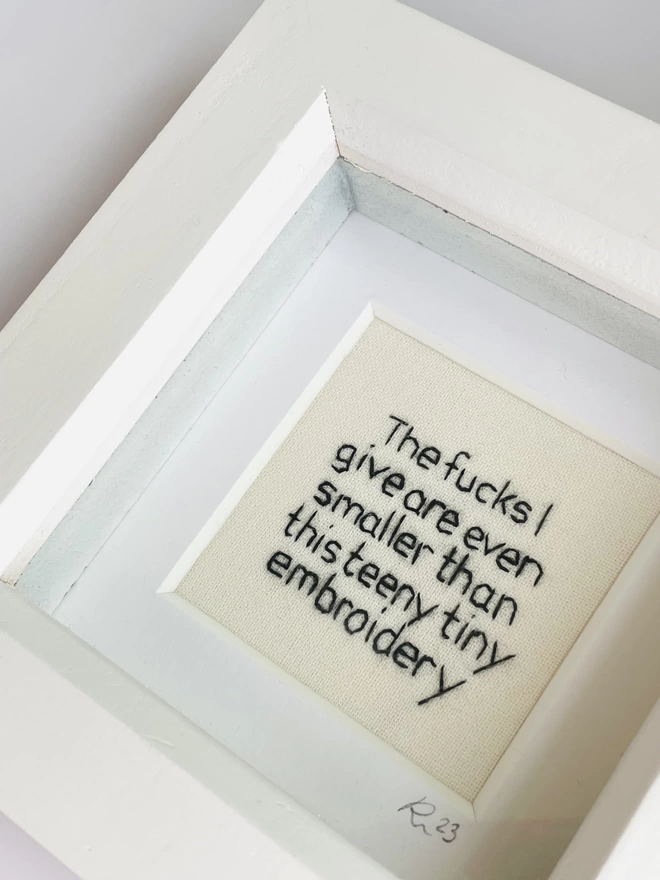 A miniature white box frame with hand embroidered black text that reads The fucks I give are even smaller than this teeny tiny embroidery