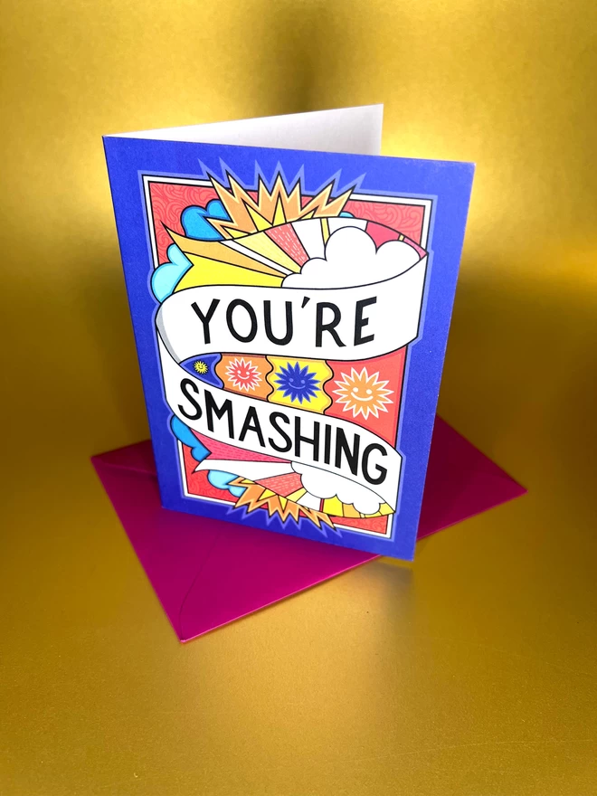 A blue greetings card featuring “You’re Smashing” written in black on a white swirl across a vibrant design of the sky featuring clouds and drawings of the sun with smiling faces, sits on a red envelope in front of a gold backdrop.