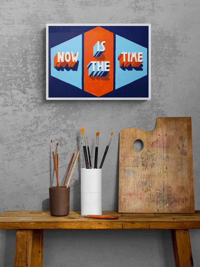 Framed art print of words Now Is The Time painted in 3d typography in orange and blue by artist Survival Techniques. Frame hangs above a table with paintbrushes and a paint palette