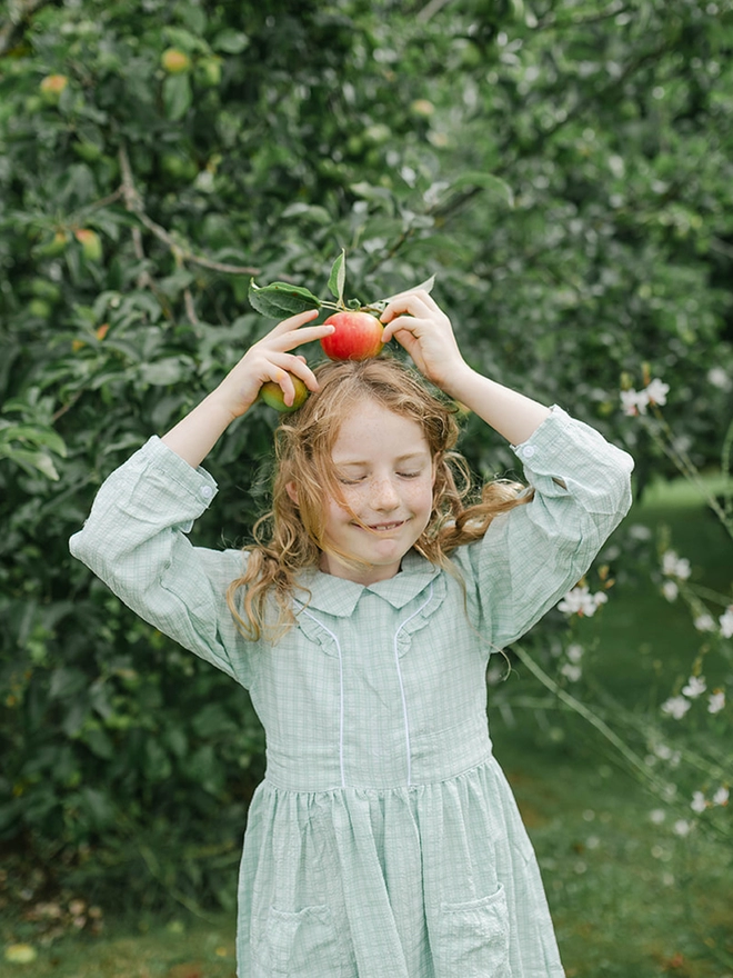 A girl in a green checked dress with long sleeves and a collar holds an apple on top of her head