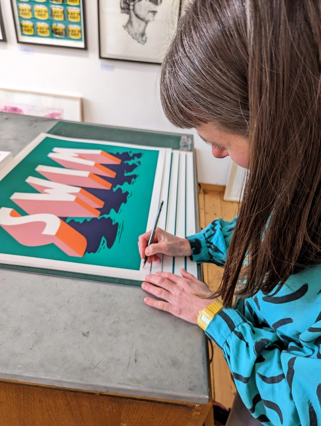 Artist Survival Techniques signing an edition of screenprints of the word SWIM in 3d typography in pink, orange and blue on a green background.