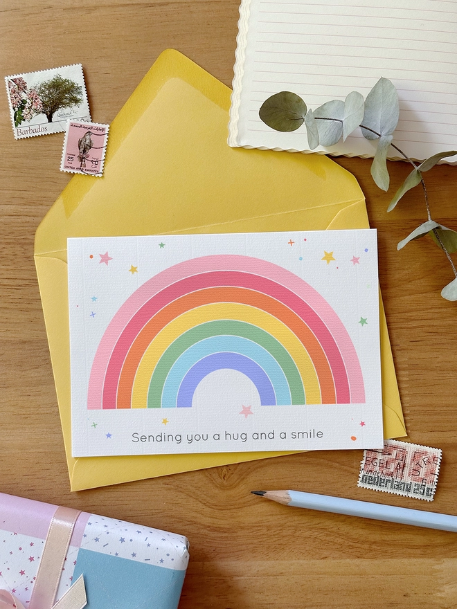 A 'sending a hug' greetings card with a pastel rainbow design lays on a yellow envelope on a wooden desk beside various stationery items.