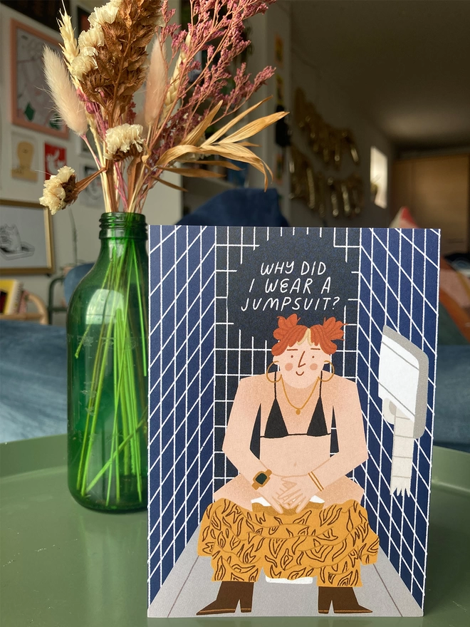 Greetings card illustrated with a semi-naked girl sat in a public toilet - questioning 'why did I wear a jumpsuit?'