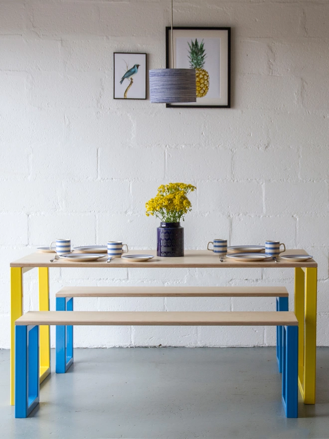 bold dining table and benches with yellow and blue boxy steel legs and birch plywood top, laid for dinner with a vase of yellow flowers in the centre