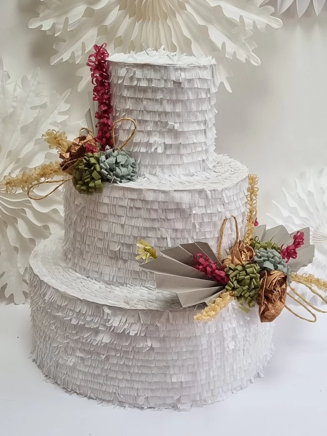 simple wedding cake pinata with dried flower style floral details on a background of white paper fans