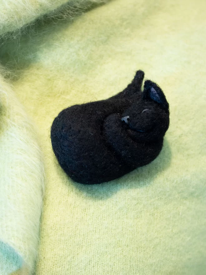 A needle feltedblack sleeping cat brooch on a green fluffy jumper 