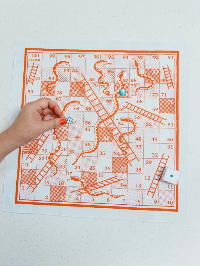 A Mr.PS orange Snakes and Ladders board game handkerchief with a blue cut out counter being played by a white hand with red nail varnish