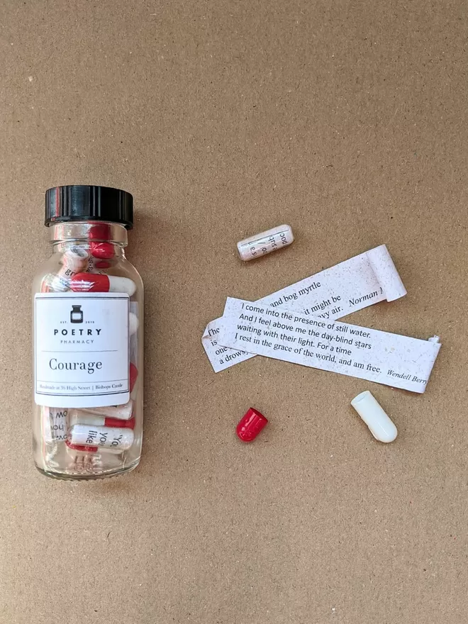 A glass bottle of Courage poetry pills alongside two capsules, one open to reveal the slips of unrolled paper with poetic quotes on it