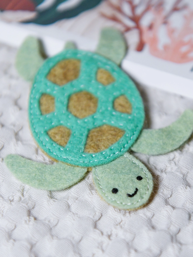 A green felt sea turtle has been sewn together with a smiling face rests on a white fabric surface.