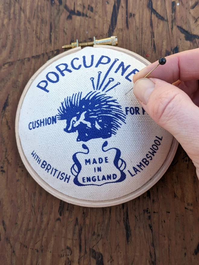 hand placing a pin in a royal blue embroidery hoop porcupine pin cushion
