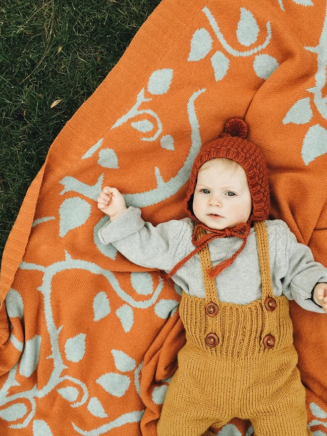 A close up of a baby lying on the rust briar rose junior blanket, the baby is wearing a cosy knitted ensemble in autumn colours which compliment the blanket.