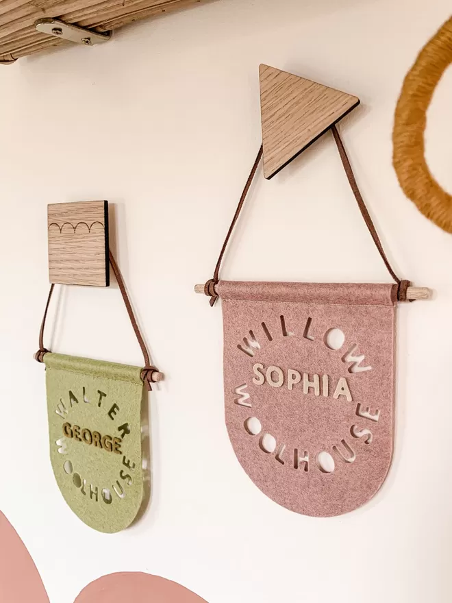 Two Name Flags on pink and green felt. Both hanging from small wooden hooks.