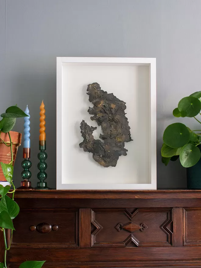 Blackend metal topographic contour map of the Peak District National Park in a white frame, sat on a wooden mantlepiece with candlesticks and plants.