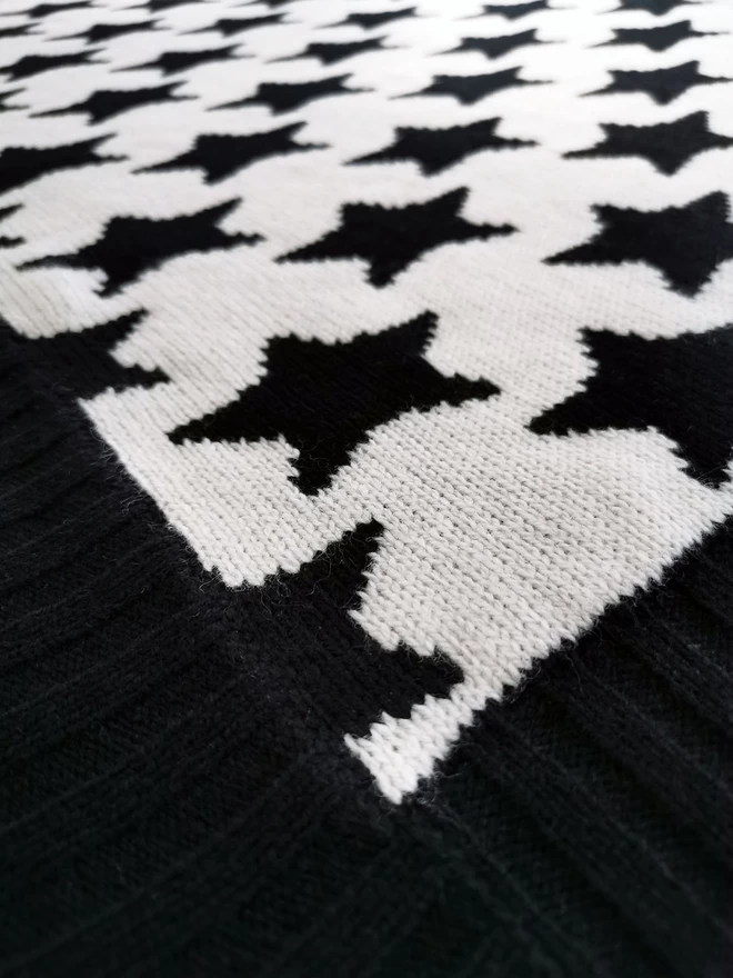 A close up of the corner of a knitted blanket showing the knit stitches, black and white star design and black ribbed trim.