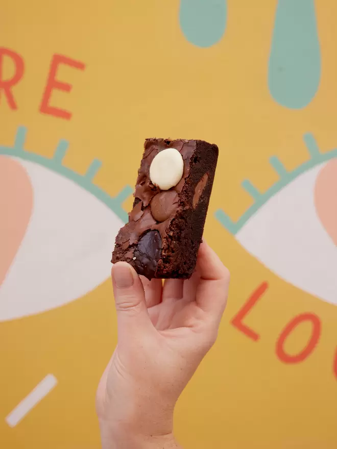 Single slice of triple chocolate button brownie being held in one hand against colourful background