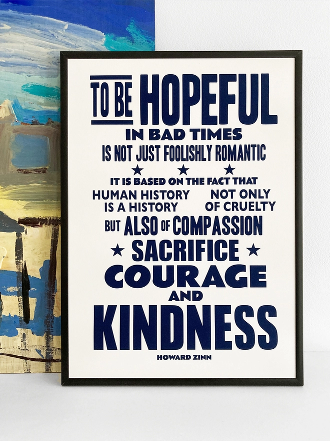 Framed typographic print of a quote by Howard Zinn. It reads "To be hopeful in bad times is not just foolishly romantic, it is based on the fact that human history is a history not only of cruelty but also of compassion, sacrifice, courage and kindness". Navy blue text on white paper.  The print rests against a blue and yellow abstract painting.