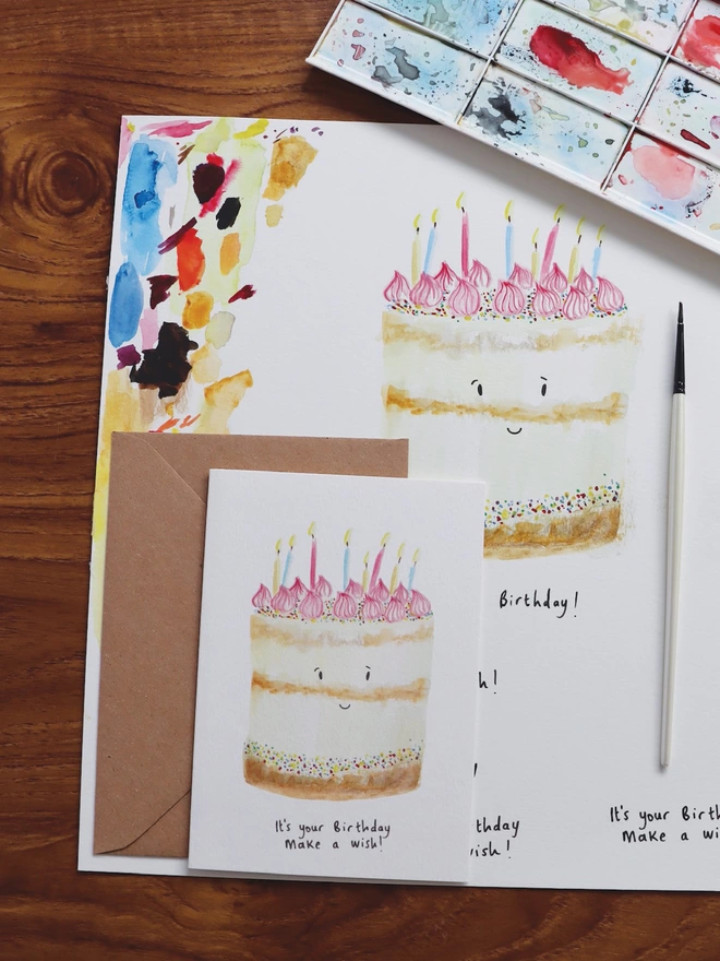 Desk Shot Of The Birthday Cake Card Sitting Along Side The Original Hand Painted Watercolour Illustration, Paintbrush and Palette
