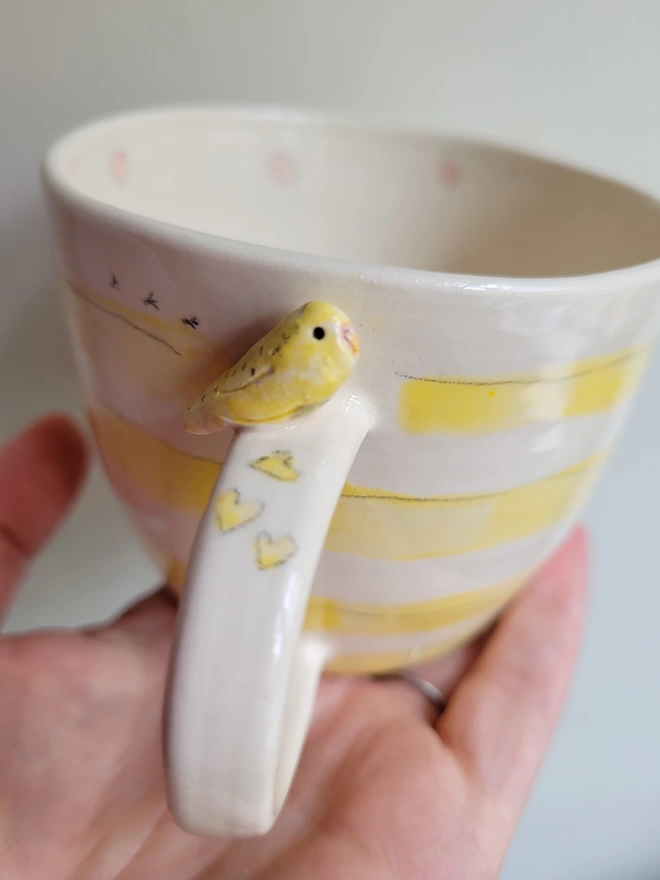 Close up of a yellow budgerigar on a ceramic stripey yellow cup with birdprints and pink dotsconbthd inside 