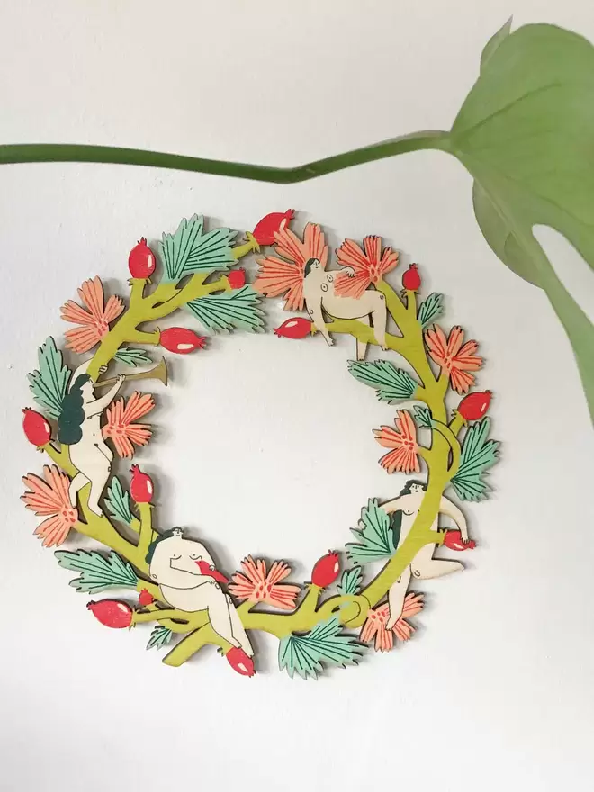 The colourful woodland wreath is hung up on a white wall, a large plant stem crosses the photo 