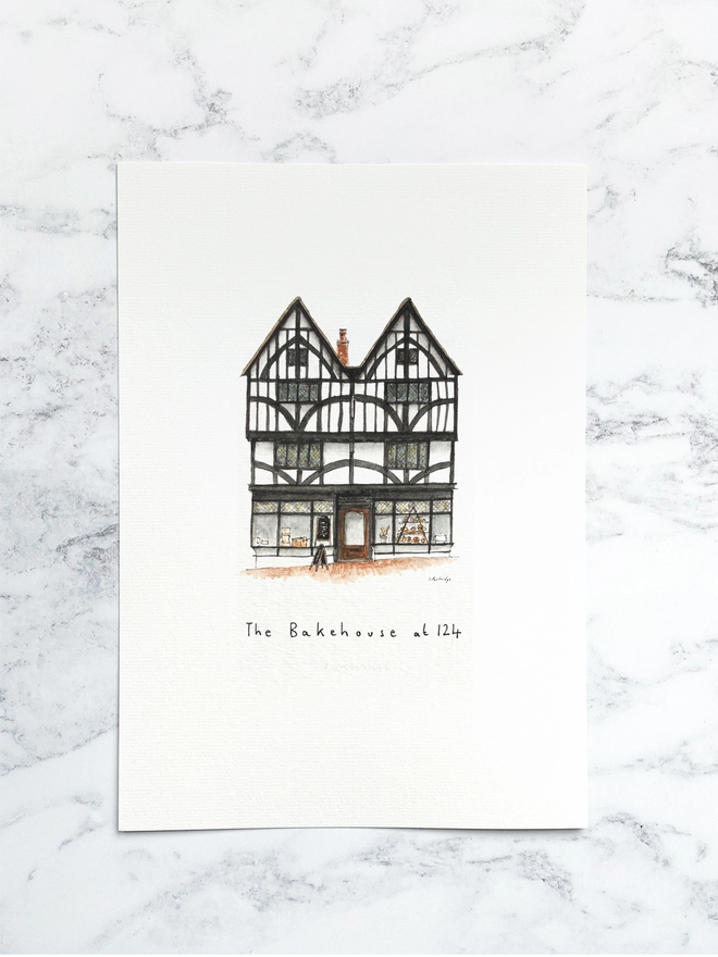 Beautiful watercolour illustration of The Bakehouse at 124, bakery in Tonbridge.  A characterful black and white tudor style three storey building with large windows on the ground floor allowing a glimpse into the bakery.The watercolour style is painted with a black pen outline and organic loose style with small details. The print is a small illustration on the centre of a white page and the paper sits on a white marble background.