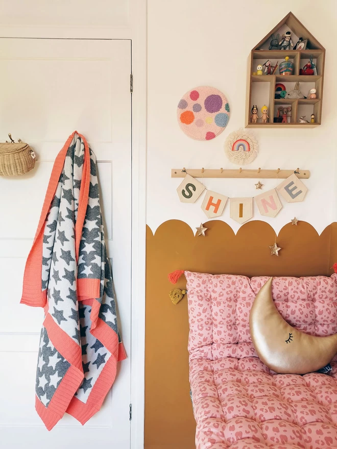 A modern childrens room with an ochre scalloped half wall. There is a cosy reading nook with a coral pink floor cushion and gold moon cushion to the right and a grey and white junior star blanket with coral pink trim hangs on a peg on the back of the door to the left. Complimentary children's decor adorns the wall.