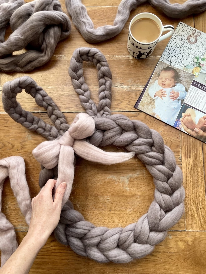 Taken from above, a soft grey merino rabbit wreath is having its light pink bow tied on an oak table, with a cup of tea and a magazine to the side. The magazine shows an image of a nursery with the same rabbit wreath hung on the wall