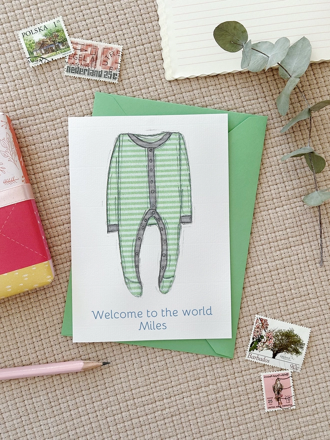 An illustrated new baby greetings card with a green onesie design lays on a green envelope beside various stationery items.