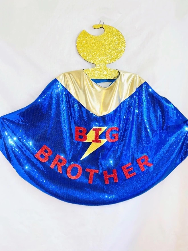 A big brother cape hanging on a hook. The cape is made from dark blue sequins and has a gold neck fastening. The text reads 'BIG BROTHER' in red and has a gold lightening bolt layered under the word 'BIG'