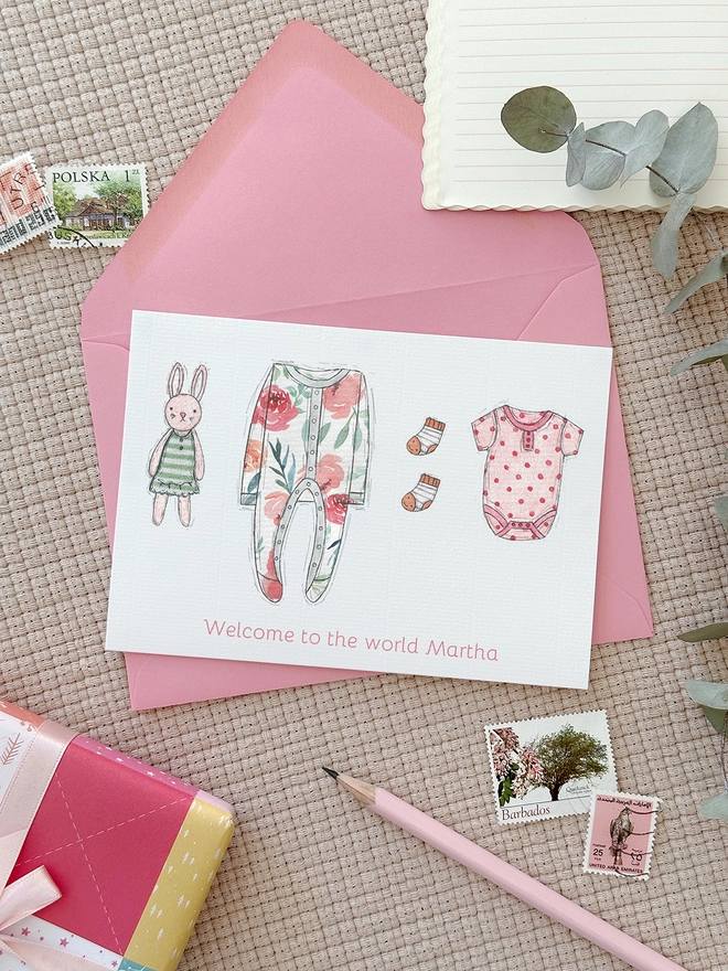 A personalised new baby greetings card with illustrated baby clothes lays on a pink envelope beside various stationery items.