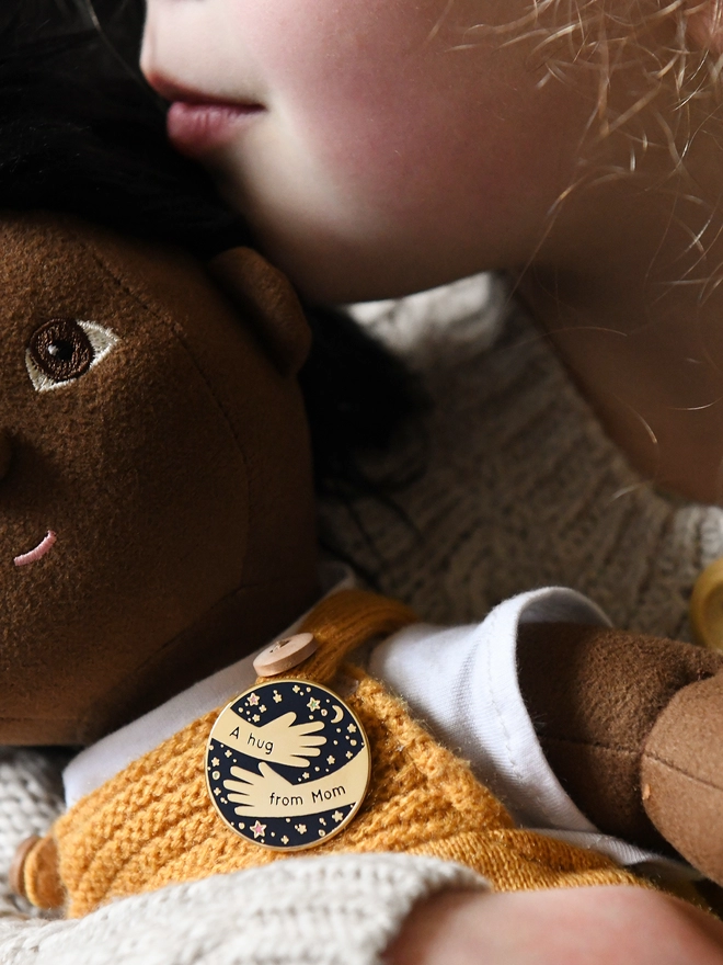 A navy blue and gold enamel pin badge with a hugging arms design and the words "A hug from Mom" is pinned to the clothes of a doll being hugged by a young child.