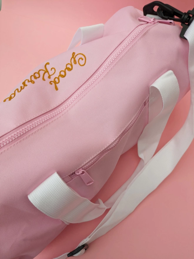 Pink Duffel Bag close up detail with white straps and handles, personalised with gold embroidery reading 'Good Karma' on a pink background showing zip detail