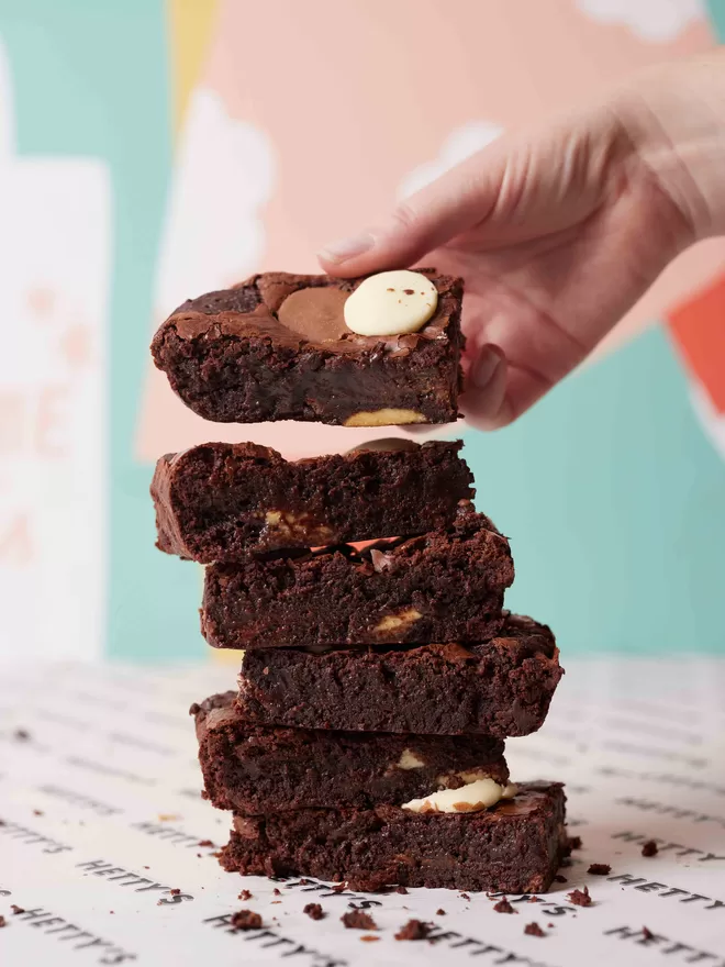 Triple chocolate brownies in a stack with a hand adding another brownie to the top for scale against a colourful background