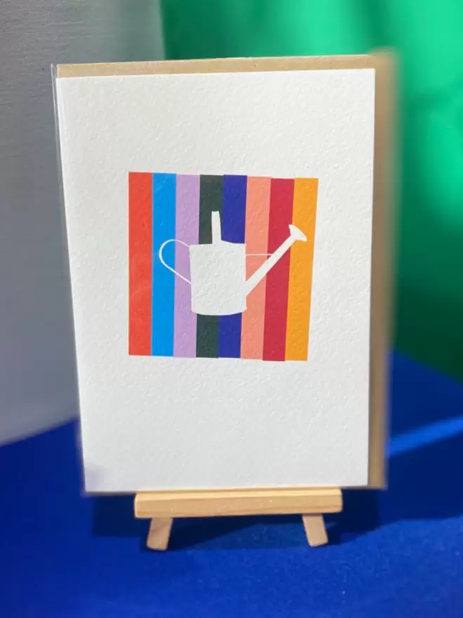 Photograph of a watering can greetings cards designed by artist, Julie-Anne featuring a colourful square of stripes and the silhouette of a white watering can over the top.